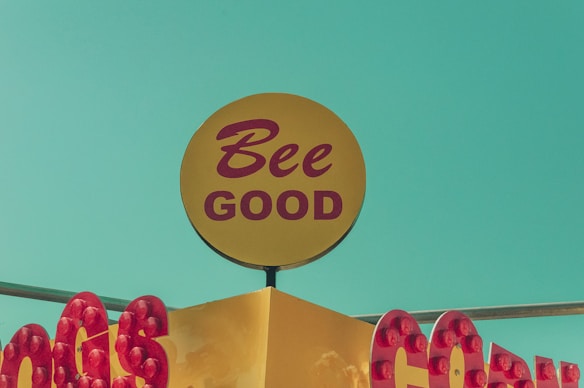 A vintage-style sign features the phrase 'Bee Good' in red letters on a yellow circular background. Red bulbs frame the signage, which sits against a bright turquoise-blue sky.