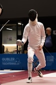 A fencer in full protective gear, including a mask and white outfit, is mid-action during a fencing match. The scene appears to be indoors with a blue advertisement panel in the background. The fencer's opponent is partially visible, and an older man in a blue shirt is watching the match.