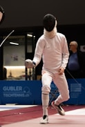 A fencer in full protective gear, including a mask and white outfit, is mid-action during a fencing match. The scene appears to be indoors with a blue advertisement panel in the background. The fencer's opponent is partially visible, and an older man in a blue shirt is watching the match.