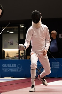 A fencer in full protective gear, including a mask and white outfit, is mid-action during a fencing match. The scene appears to be indoors with a blue advertisement panel in the background. The fencer's opponent is partially visible, and an older man in a blue shirt is watching the match.
