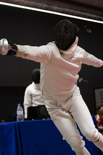 A fencer in protective gear lunges forward with a foil in hand, showcasing a dynamic pose during a match. Another person, also in fencing attire, is visible in the background near a table with a water bottle and equipment.