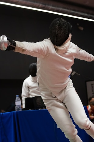 A fencer in protective gear lunges forward with a foil in hand, showcasing a dynamic pose during a match. Another person, also in fencing attire, is visible in the background near a table with a water bottle and equipment.