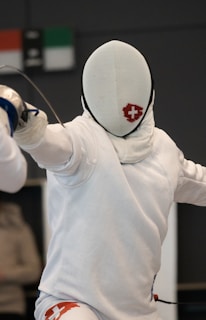 A fencer dressed in white protective gear, including a mask, is positioned in a stance, holding a fencing sword. The mask features a red cross logo, and the fencer's movement suggests engagement in a bout. Background elements are blurred, focusing attention on the fencer.