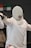 A fencer dressed in white protective gear, including a mask, is positioned in a stance, holding a fencing sword. The mask features a red cross logo, and the fencer's movement suggests engagement in a bout. Background elements are blurred, focusing attention on the fencer.