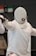 A fencer dressed in white protective gear, including a mask, is positioned in a stance, holding a fencing sword. The mask features a red cross logo, and the fencer's movement suggests engagement in a bout. Background elements are blurred, focusing attention on the fencer.