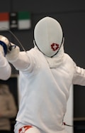 A fencer dressed in white protective gear, including a mask, is positioned in a stance, holding a fencing sword. The mask features a red cross logo, and the fencer's movement suggests engagement in a bout. Background elements are blurred, focusing attention on the fencer.