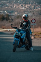 A person wearing a black leather jacket and a helmet with a unique design is sitting on a vibrant blue motorcycle, parked on a road. In the background, there are blurred views of a cityscape and trees, with a road sign visible nearby.