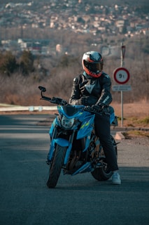 A person wearing a black leather jacket and a helmet with a unique design is sitting on a vibrant blue motorcycle, parked on a road. In the background, there are blurred views of a cityscape and trees, with a road sign visible nearby.