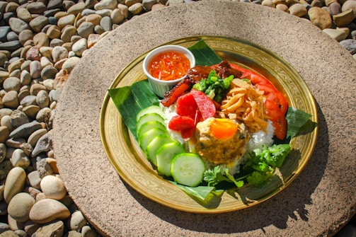 A vibrant dish arranged on a decorative golden plate resting on a textured stone surface surrounded by small pebbles. The dish includes white rice topped with various colorful ingredients such as slices of cucumber, tomato, red sausage, caramelized onions, a yellow sauce with an egg yolk, and garnished with cilantro and green onions. A small bowl of red dipping sauce accompanies the meal.