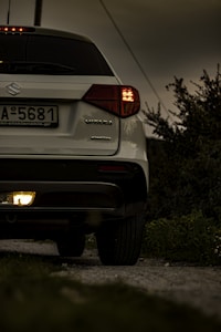 A white Suzuki Vitara is parked on a gravel path, surrounded by greenery under a cloudy sky. The car's rear lights are illuminated, suggesting it might be stationary but ready to move. The license plate is visible, and the badge indicates it's an AllGrip model.
