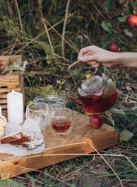 A tranquil outdoor setting featuring a hand pouring tea from a glass teapot into a cup on a wooden tray. The tray also holds slices of cake on parchment paper, two additional glass cups, and a lit white candle. Surrounding this scene are lush green plants and ripe red apples on the ground.