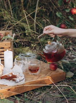 A tranquil outdoor setting featuring a hand pouring tea from a glass teapot into a cup on a wooden tray. The tray also holds slices of cake on parchment paper, two additional glass cups, and a lit white candle. Surrounding this scene are lush green plants and ripe red apples on the ground.