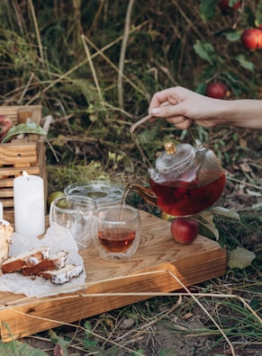 A tranquil outdoor setting featuring a hand pouring tea from a glass teapot into a cup on a wooden tray. The tray also holds slices of cake on parchment paper, two additional glass cups, and a lit white candle. Surrounding this scene are lush green plants and ripe red apples on the ground.