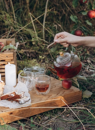 A tranquil outdoor setting featuring a hand pouring tea from a glass teapot into a cup on a wooden tray. The tray also holds slices of cake on parchment paper, two additional glass cups, and a lit white candle. Surrounding this scene are lush green plants and ripe red apples on the ground.