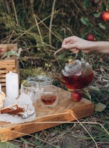 A tranquil outdoor setting featuring a hand pouring tea from a glass teapot into a cup on a wooden tray. The tray also holds slices of cake on parchment paper, two additional glass cups, and a lit white candle. Surrounding this scene are lush green plants and ripe red apples on the ground.