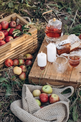 A rustic outdoor setup featuring a wooden crate filled with red apples, a wooden table with lit white candles, a glass teapot containing tea, slices of cake on crumpled paper, and woven cloth holding more apples. The setting is surrounded by grass, creating a cozy and inviting autumnal ambiance.