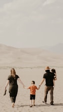 Four people, presumably a family, walk together in a sandy desert landscape. The adults, one wearing a hat, hold the hands of two children, one of whom is being carried. The background features sandy dunes and a mountainous horizon under a cloudy sky.