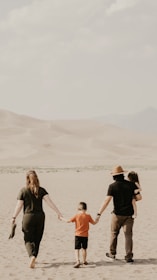 Four people, presumably a family, walk together in a sandy desert landscape. The adults, one wearing a hat, hold the hands of two children, one of whom is being carried. The background features sandy dunes and a mountainous horizon under a cloudy sky.