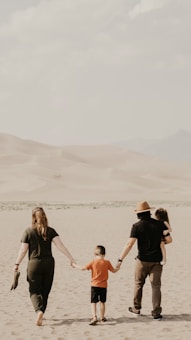 Four people, presumably a family, walk together in a sandy desert landscape. The adults, one wearing a hat, hold the hands of two children, one of whom is being carried. The background features sandy dunes and a mountainous horizon under a cloudy sky.