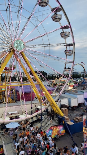 A crowded amusement park features a large Ferris wheel with bright lights. Below, numerous people gather in lines, suggesting a lively atmosphere. Various rides and stalls can be seen in the background, adding to the vibrant festival setting.