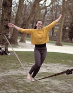 A person confidently balances on a slackline in a park setting, surrounded by trees. They appear to be enjoying the activity, wearing a vibrant yellow sweater and dark pants, with arms outstretched for balance. The background features people sitting and the park environment.
