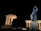 A large classical statue is illuminated against the night sky, situated beside a temple-like structure with columns. The statue depicts a robed figure with a lion in a dynamic, heroic pose.