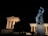 A large classical statue is illuminated against the night sky, situated beside a temple-like structure with columns. The statue depicts a robed figure with a lion in a dynamic, heroic pose.