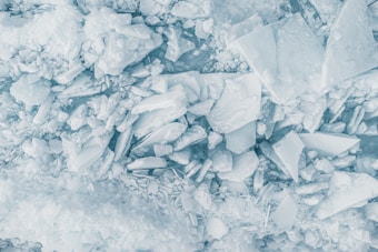 A series of fractured and stacked ice sheets in various shapes and sizes create a complex pattern. The ice pieces have a translucent quality and pale blue hue, casting soft shadows. Some ice fragments are large and angular, while others are smaller and more rounded, forming an uneven texture.