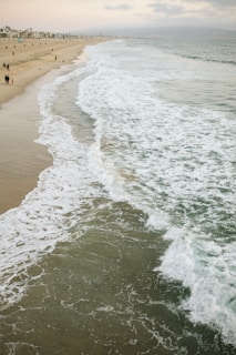 A serene beach scene with gentle waves rolling onto the sandy shore. Sparse clouds linger in the sky, while several small groups of people stroll along the coast. The horizon stretches far, with numerous beachfront properties lining the distant shoreline.