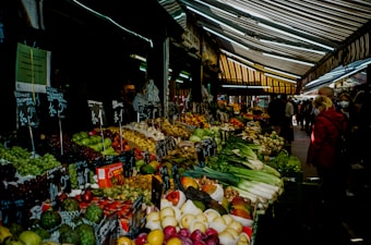 A bustling outdoor market stall is filled with a variety of fresh fruits and vegetables. The display includes green apples, grapes, artichokes, and leafy vegetables, neatly organized with handwritten price tags. People are seen browsing and wearing coats, suggesting a cool weather. The scene is sheltered by colorful striped awnings that add a vibrant contrast.