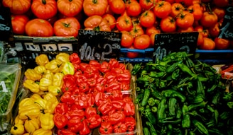 A vibrant display of fresh vegetables at a market stall featuring an assortment of produce. The top row consists of ripe red tomatoes with green stems, arranged neatly in crates. Below, there are three sections of colorful peppers: bright yellow bell peppers on the left, red chili peppers in the center, and green chili peppers on the right. Prices are displayed on small black chalkboards.