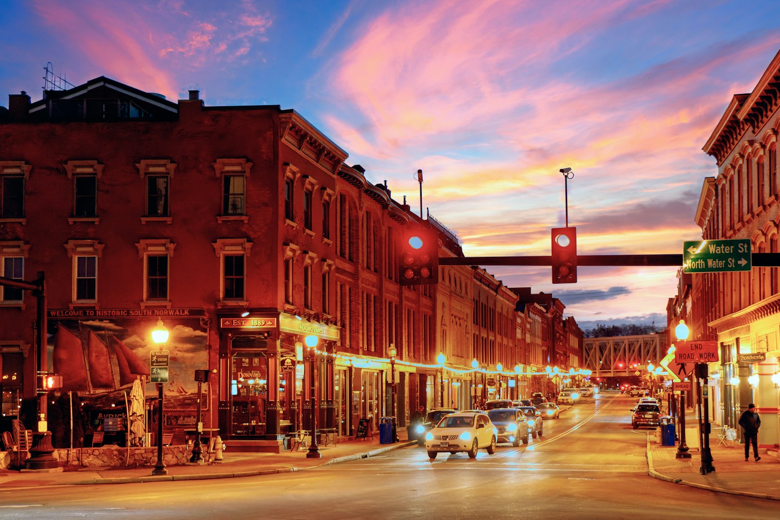 A downtown main street at sunset with cars, traffic light, and brick storefronts