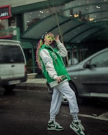 A person with colorful braided hair is wearing a green and white varsity jacket and matching sneakers. They have vibrant green sunglasses and are holding a smartphone. The setting appears to be an urban street with parked cars and a building in the background.