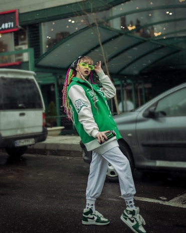 A person with colorful braided hair is wearing a green and white varsity jacket and matching sneakers. They have vibrant green sunglasses and are holding a smartphone. The setting appears to be an urban street with parked cars and a building in the background.