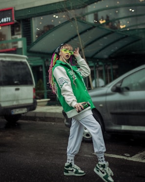 A person with colorful braided hair is wearing a green and white varsity jacket and matching sneakers. They have vibrant green sunglasses and are holding a smartphone. The setting appears to be an urban street with parked cars and a building in the background.