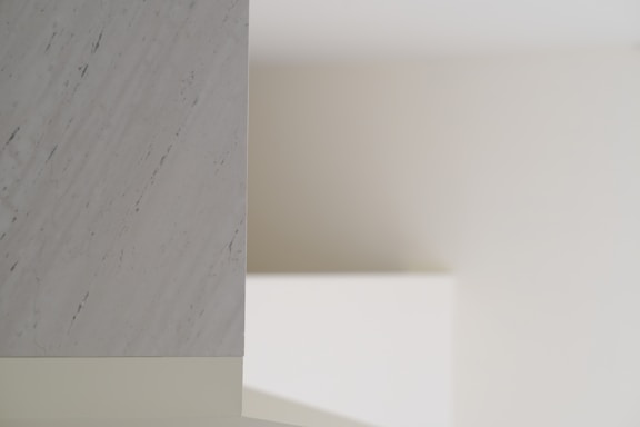 A minimalist interior scene with soft lighting, featuring a smooth marble surface on the left with subtle gray veining. The background consists of plain, neutral-colored walls creating shadows and depth.