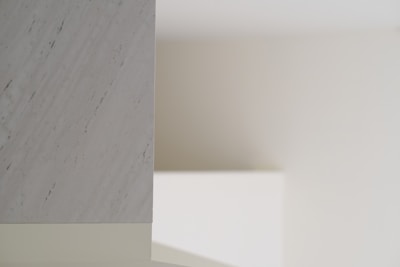 A minimalist interior scene with soft lighting, featuring a smooth marble surface on the left with subtle gray veining. The background consists of plain, neutral-colored walls creating shadows and depth.