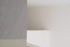 A minimalist interior scene with soft lighting, featuring a smooth marble surface on the left with subtle gray veining. The background consists of plain, neutral-colored walls creating shadows and depth.