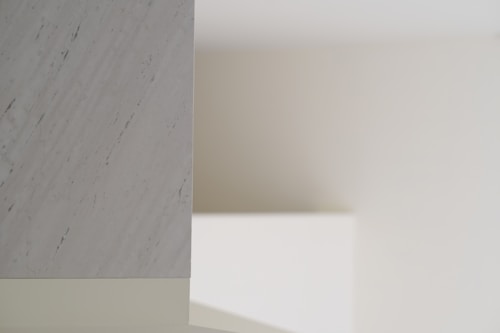 A minimalist interior scene with soft lighting, featuring a smooth marble surface on the left with subtle gray veining. The background consists of plain, neutral-colored walls creating shadows and depth.