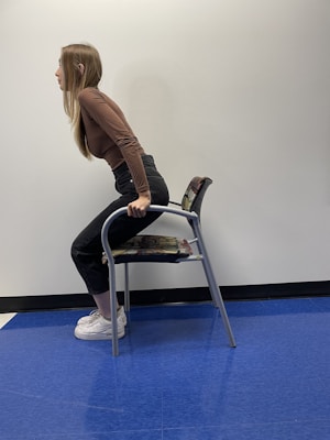 A person with long blonde hair is in a standing-sit position supported by a chair. They are wearing a brown long-sleeve top, black high-waisted pants, and white sneakers. The chair is metallic with a patterned cushion. The floor is covered with blue tiles, and the background wall is plain and light-colored.
