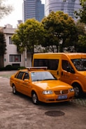 A yellow taxi is parked in front of a larger yellow van on an urban street. Several trees and modern high-rise buildings with glass windows are visible in the background.