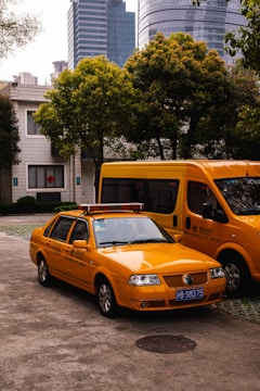 A yellow taxi is parked in front of a larger yellow van on an urban street. Several trees and modern high-rise buildings with glass windows are visible in the background.