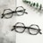 Two pairs of hexagonal eyeglasses with thick, patterned frames are placed on a textured gray and white surface. A sprig of green eucalyptus leaves lies next to the glasses, adding an organic element to the scene.