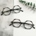Two pairs of hexagonal eyeglasses with thick, patterned frames are placed on a textured gray and white surface. A sprig of green eucalyptus leaves lies next to the glasses, adding an organic element to the scene.