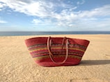 A vibrant, multicolored straw bag rests on a sandy beach under a blue sky with scattered clouds. The ocean is visible in the distance, creating a serene atmosphere.