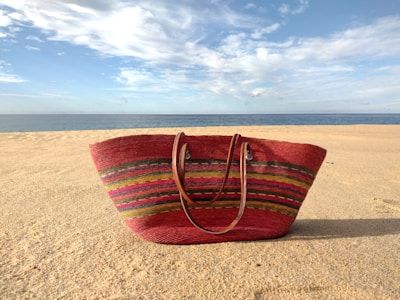 A vibrant, multicolored straw bag rests on a sandy beach under a blue sky with scattered clouds. The ocean is visible in the distance, creating a serene atmosphere.