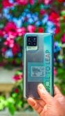 A hand holds a smartphone with a quad-camera setup, labeled with 'Dare to Leap.' The background consists of vibrant, blurred flowers in shades of pink, red, and blue, contrasting with the phone's sleek design.