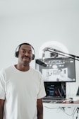 A person wearing a white t-shirt and headphones is standing in front of a microphone. Behind him, there is a computer setup with a screen displaying a digital interface. The setting appears to be a recording or podcasting space with a clean, minimalistic design.
