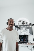 A person wearing a white t-shirt and headphones is standing in front of a microphone. Behind him, there is a computer setup with a screen displaying a digital interface. The setting appears to be a recording or podcasting space with a clean, minimalistic design.
