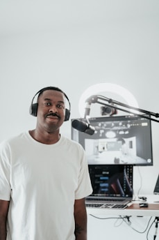 A person wearing a white t-shirt and headphones is standing in front of a microphone. Behind him, there is a computer setup with a screen displaying a digital interface. The setting appears to be a recording or podcasting space with a clean, minimalistic design.
